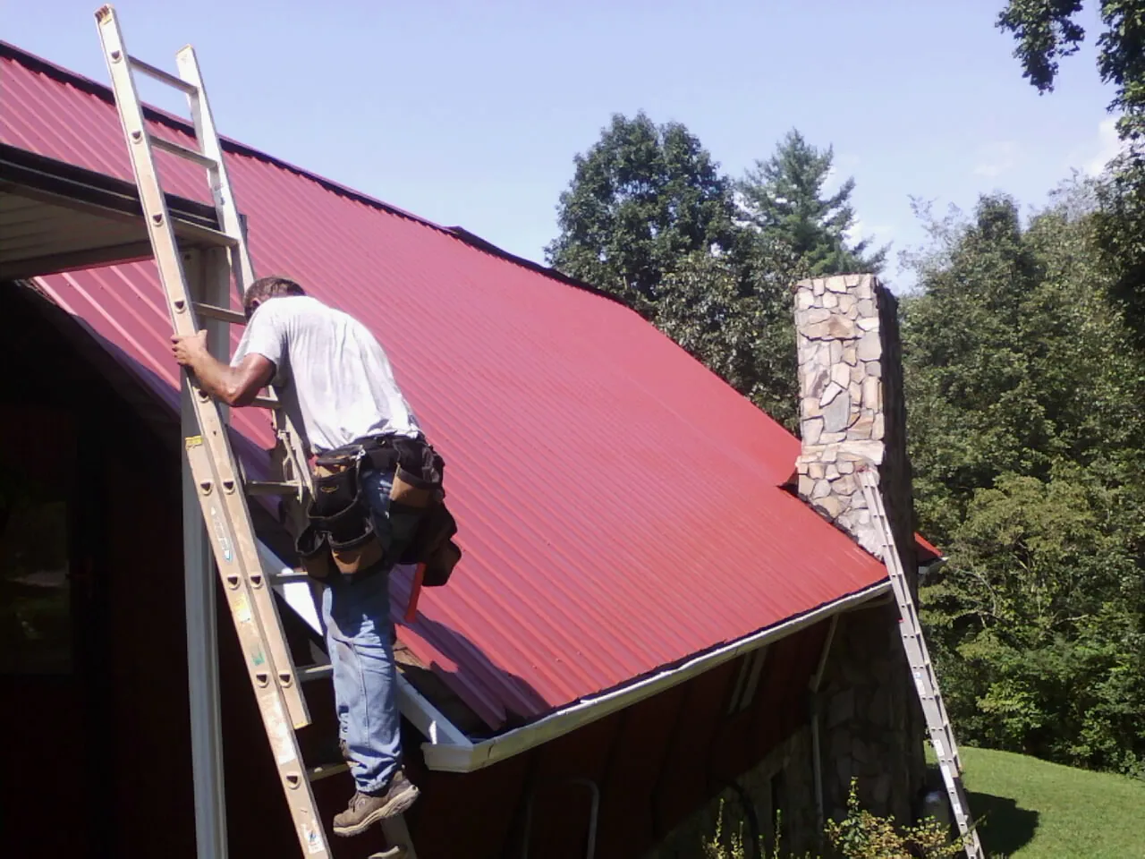 Roofer on a ladder beside a red metal roof and stone chimney — Soffit & Fascia Repair by Roofing Brothers in Oostburg